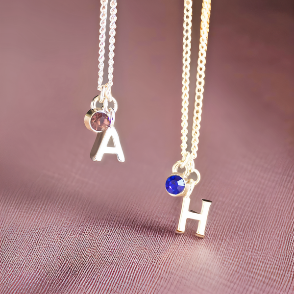 image shows two children's silver necklaces with personalised initial and birthstone charms on a pink fabric background