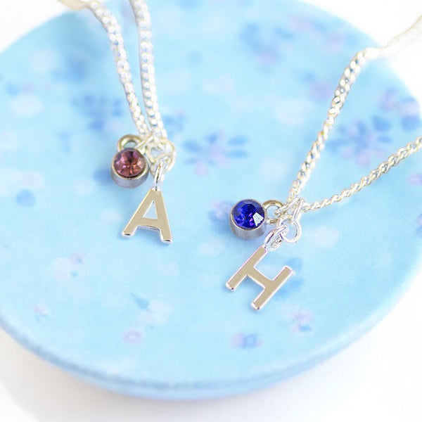 Image shows two child's silver initial necklaces with birthstone detail on a blue floral trinket dish