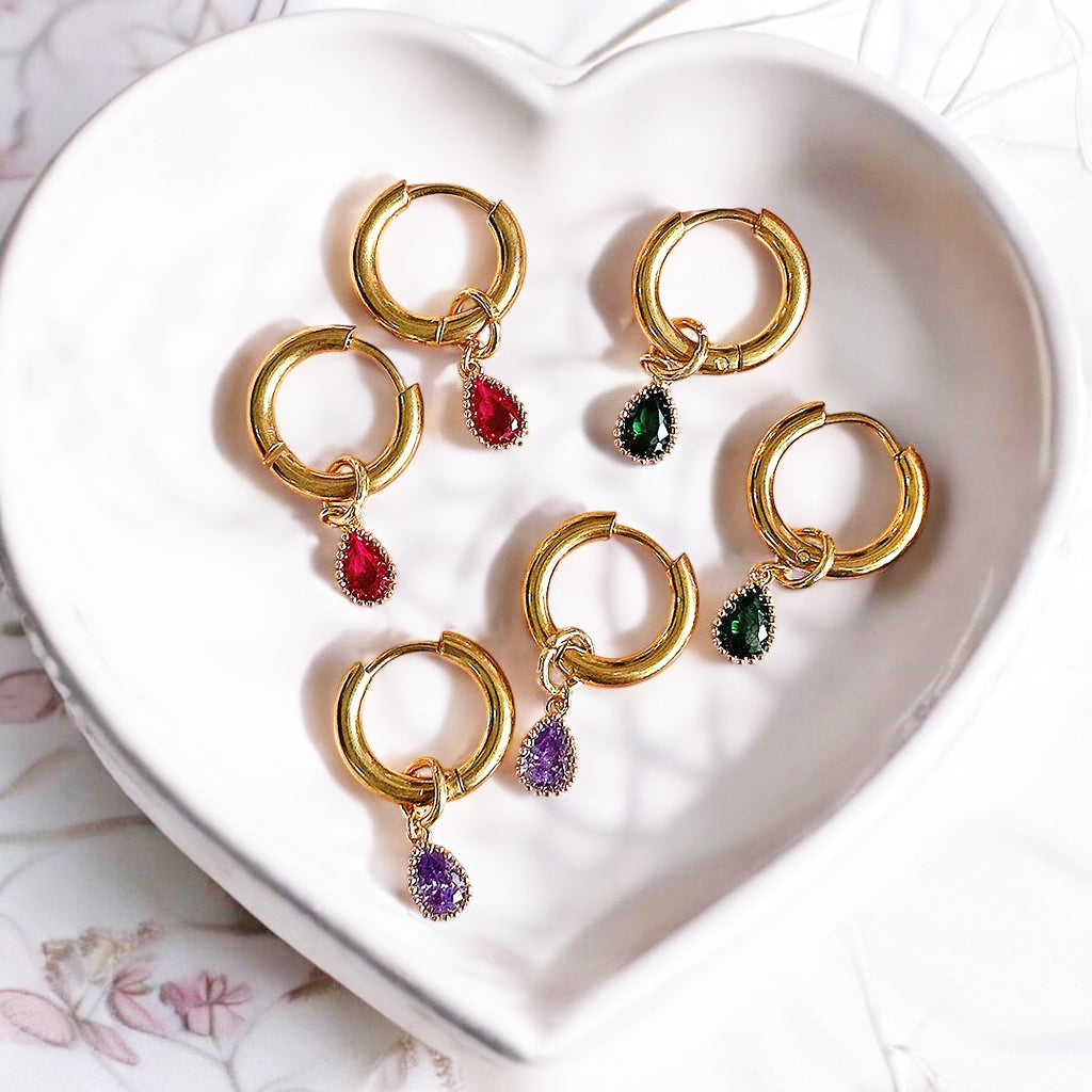 Image shows a selection of teardrop shaped birthstone earrings on gold hoops in a heart shaoped trinket dish