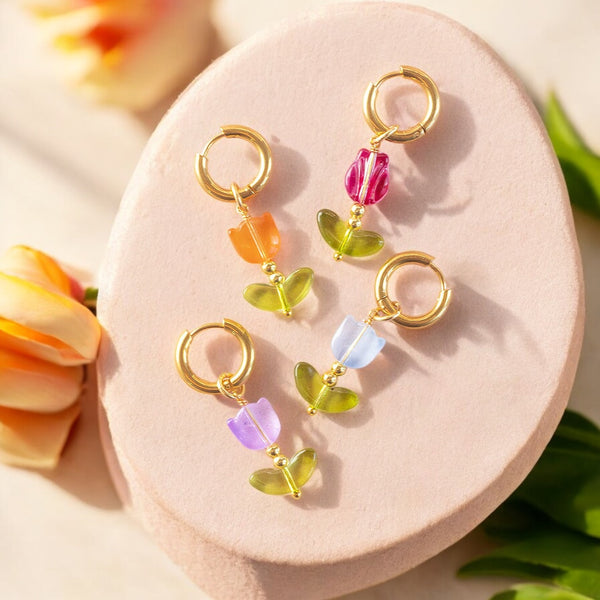 Image shows gold hoop glass tulip earrings on a peach concrete background with flowers scattered around it