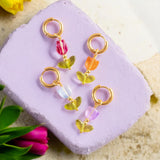 Image shows gold hoop glass tulip earrings on a lilac concrete background with flowers scattered around it