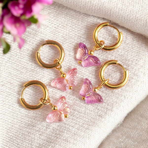 Image shows gold hoop earrings with glass butterfly charms in pink and purple shades on a neutral background with flowers