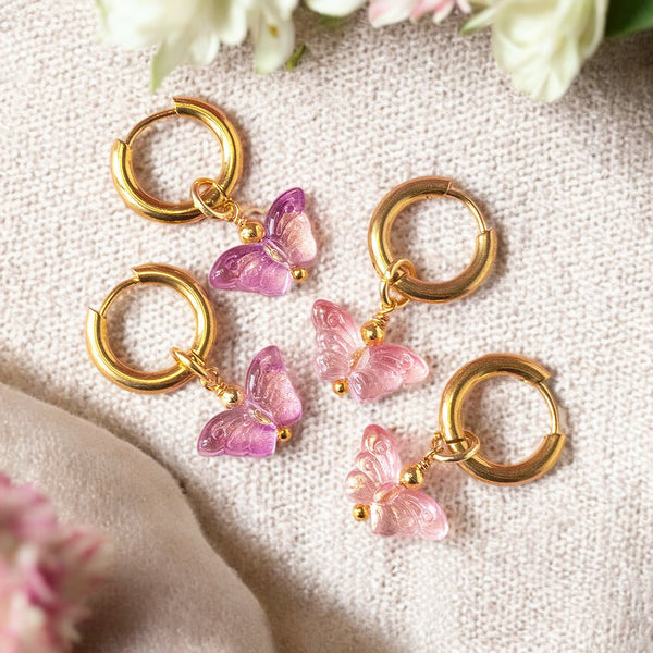 Image shows gold hoop earrings with glass butterfly charms in pink and purple shades on a neutral background with flowers