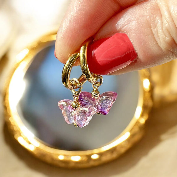 Image shows a hand holding gold hoop earrings with glass butterfly detail