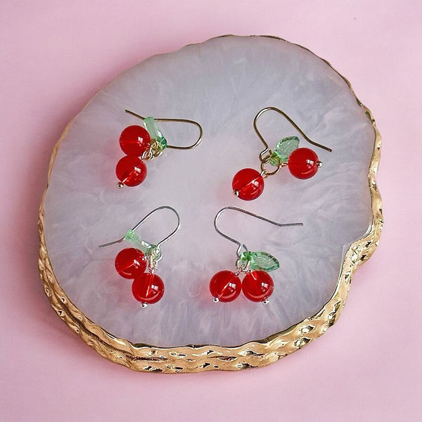 Image shows a pair of gold and a pair of silver handmade glass cherry earring on a gold edged coaster