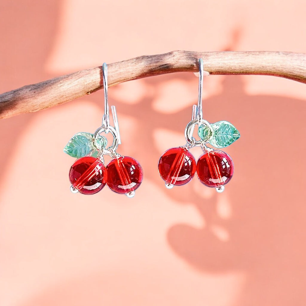 Image shows a pair of handmade glass cherry earrings hanging from a twig against a peach shadowy background