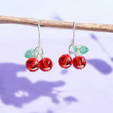 Image shows a pair of handmade glass cherry earrings hanging from a twig against a lilacshadowy background