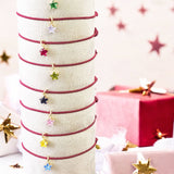 image shows a stack of maroon cord friendship bracelets with birthstone star charms in front of a pink festive backdrop