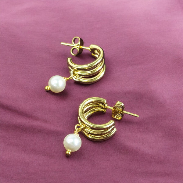 Image shows a pair of triple hoop gold plated earrings with pearl detail on a deep pink background