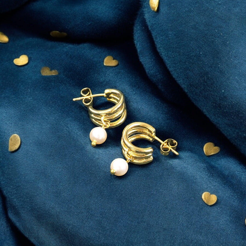 Image shows a pair of triple hoop gold plated earrings with pearl drop detail on a deep blue background with scattered gold heart