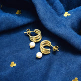 Image shows a pair of triple hoop gold plated earrings with pearl detail on a deep blue fabric background