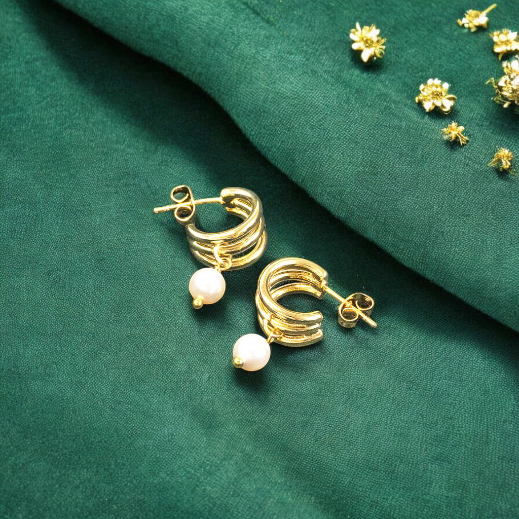 Image shows a pair of triple hoop gold plated earrings with pearl detail on a deep green background with scattered go9ld flowers