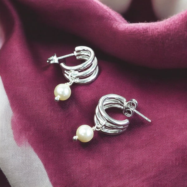 image shows a pair of triple hoop silver earrings with pearl charm detail on a maroon fabric background