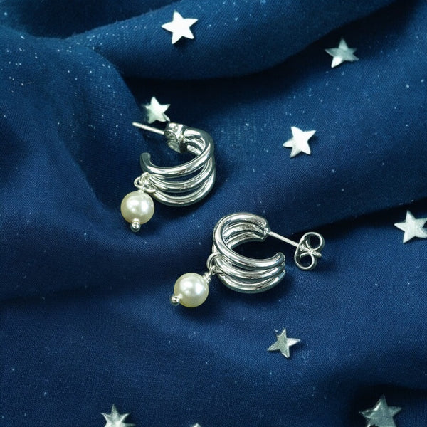 image shows a pair of triple hoop silver earrings with pearl charm detail on a deep blue fabric background with scattered silver stars