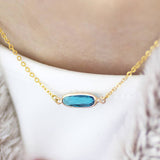 Image shows a woman in a white teeshirt wearing a blue topaz December oblong birthstone necklace