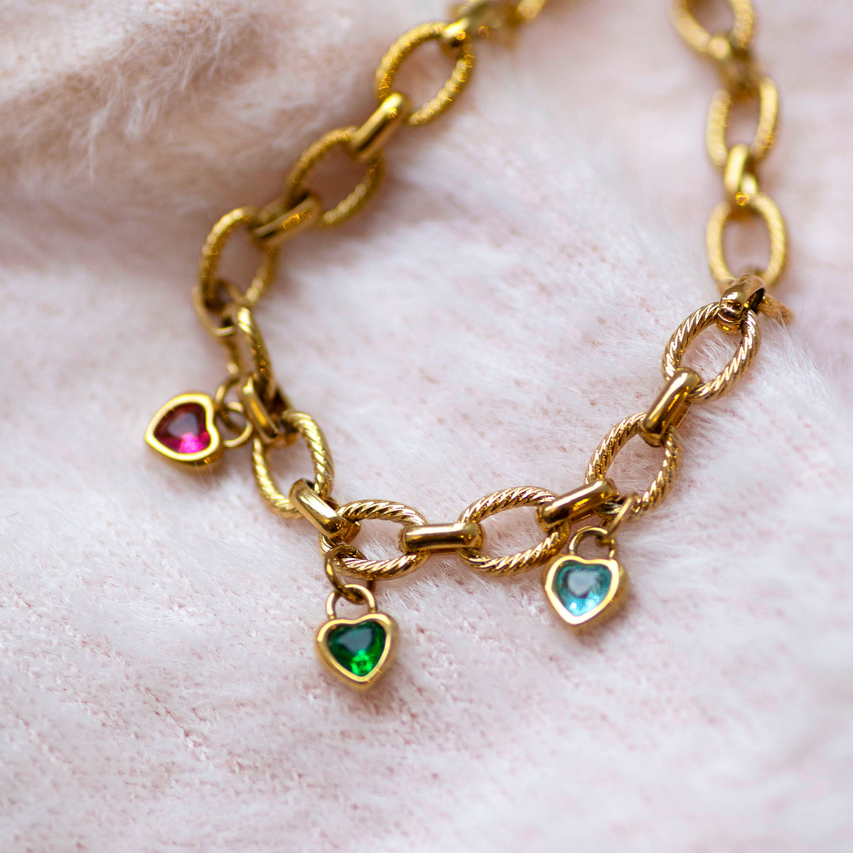 Gold chain bracelet with heart-shaped birthstone charms on a light pink fabric background