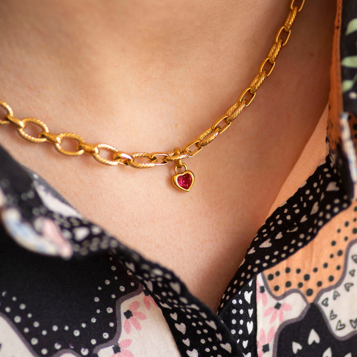 Gold chain charm necklace with a heart-shaped pendant worn by a person in a patterned top.