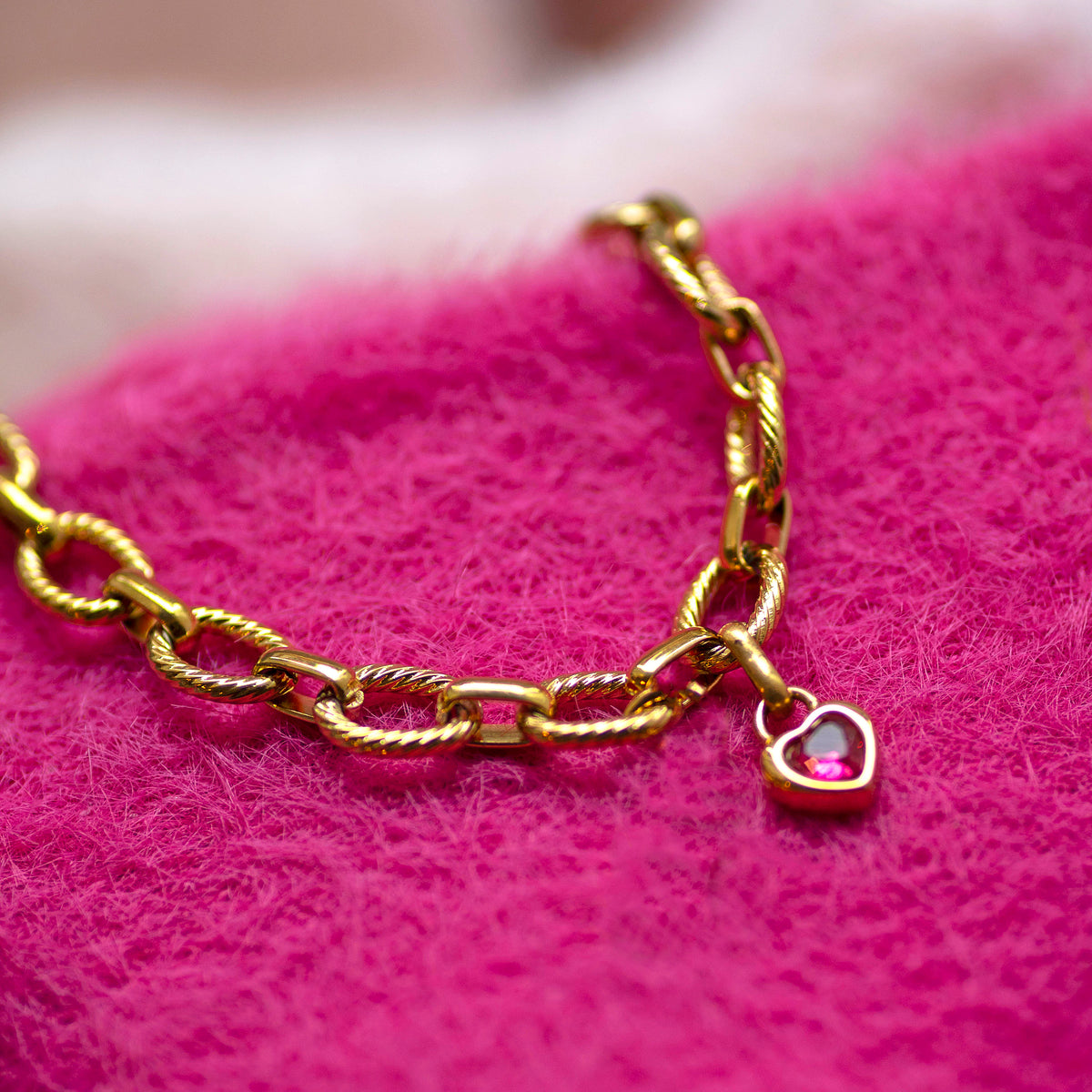Gold chain bracelet with textured oval links and a small pink heart charm on a bright pink fabric background.