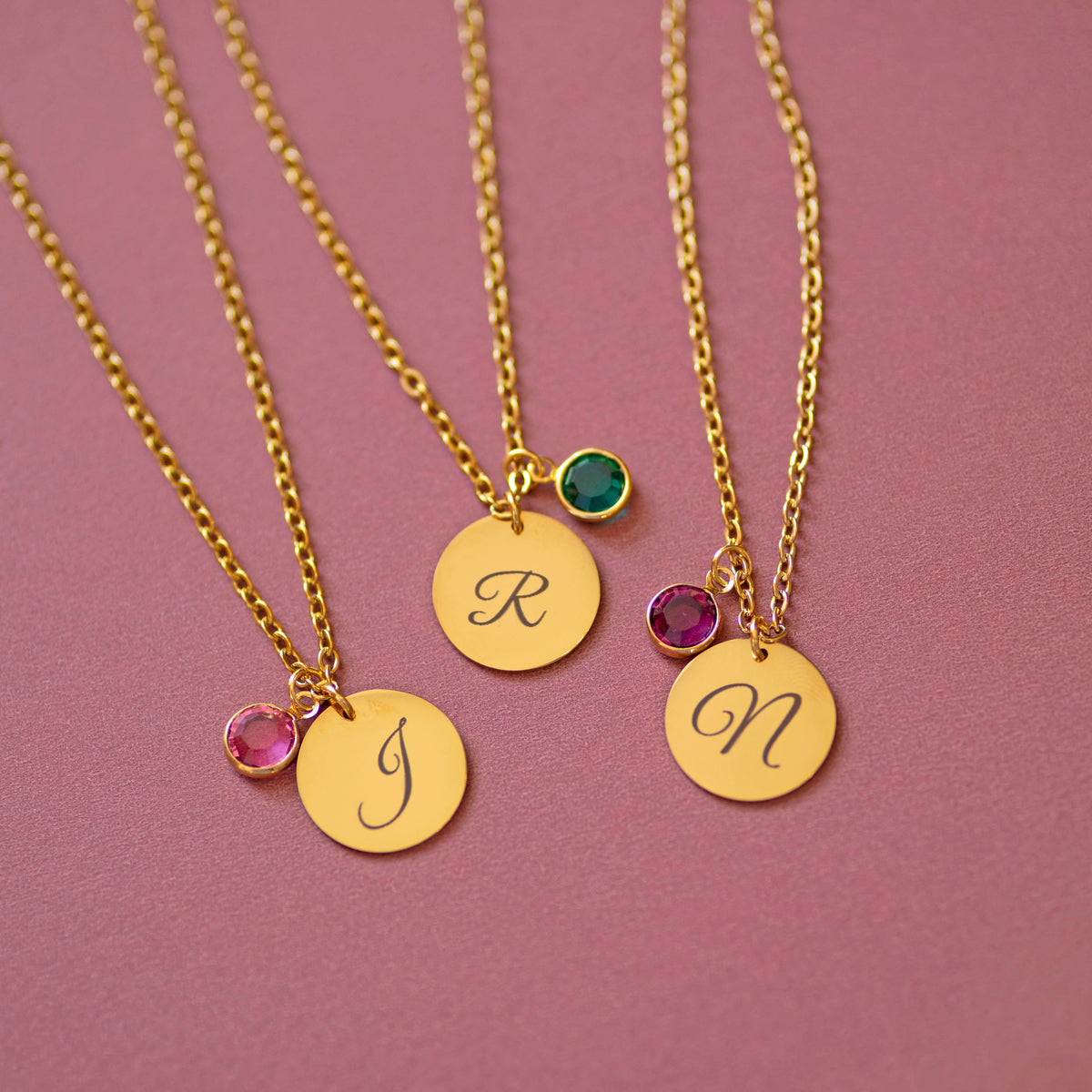 Gold script initial birthstone charm necklaces with engraved letters J, R and N, each paired with a coloured birthstone charm on a soft pink background.