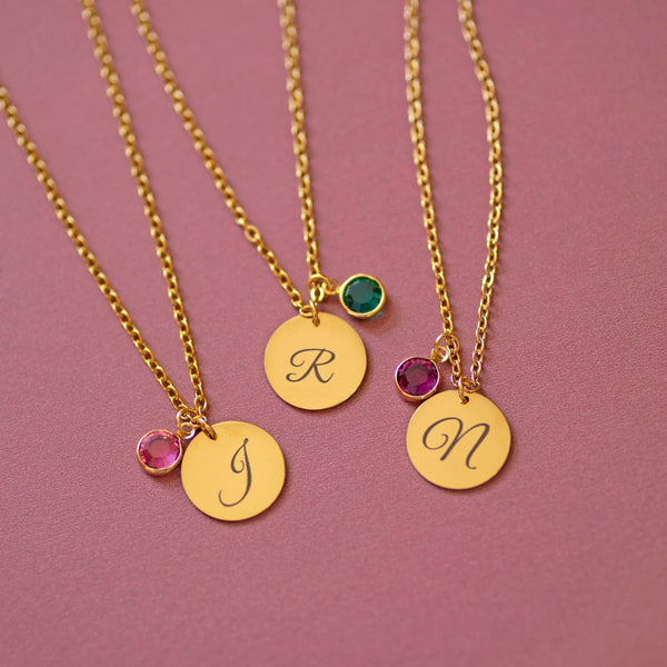 Gold script initial birthstone charm necklaces with engraved letters J, R and N, each paired with a coloured birthstone charm on a soft pink background.