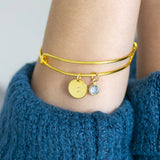 image shows a woman in a blue jumper wearing a gold adjustable bangle personalised with an initial S and march aquamarine birthstone charm