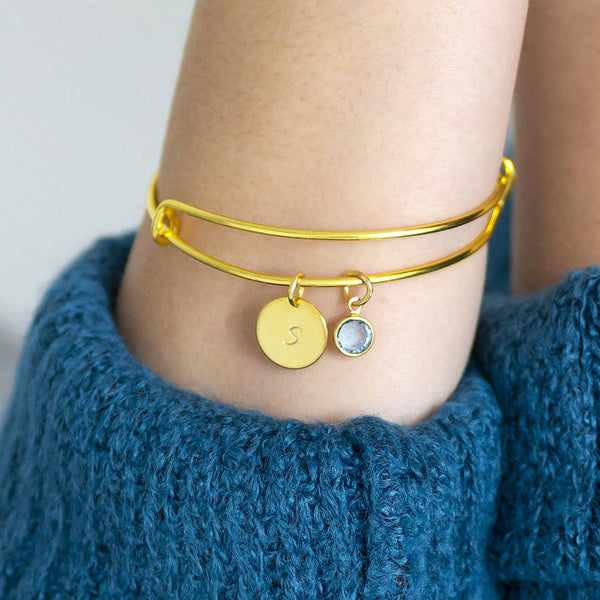 image shows a woman in a blue jumper wearing a gold adjustable bangle personalised with an initial S and march aquamarine birthstone charm