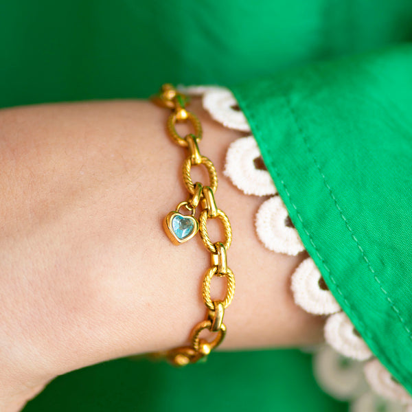 Gold link bracelet with a blue heart charm worn on a wrist, styled with a green sleeve featuring white lace trim.