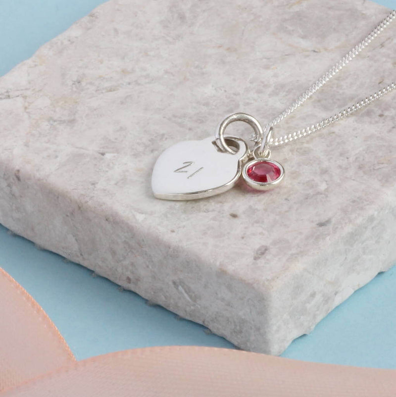 Image shows sterling silver heart charm necklace featuring a '21' engraved heart and October roseSwarovski crystal birthstone. Necklace i on a grey backdrop