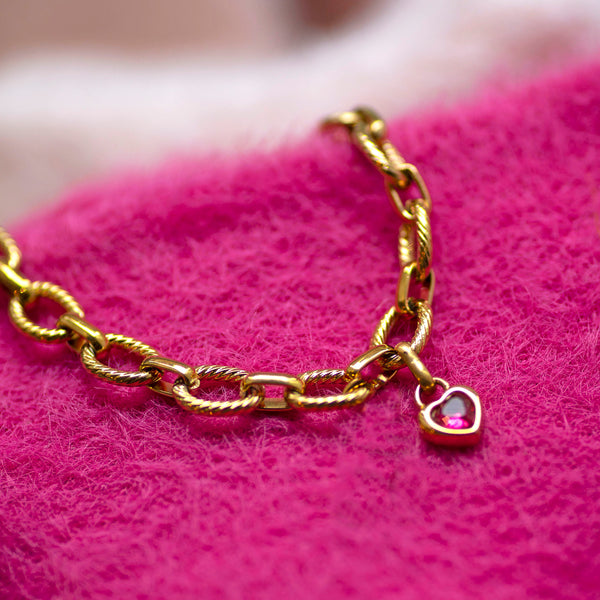 Chunky gold chain bracelet with a heart-shaped charm on a pink textured surface