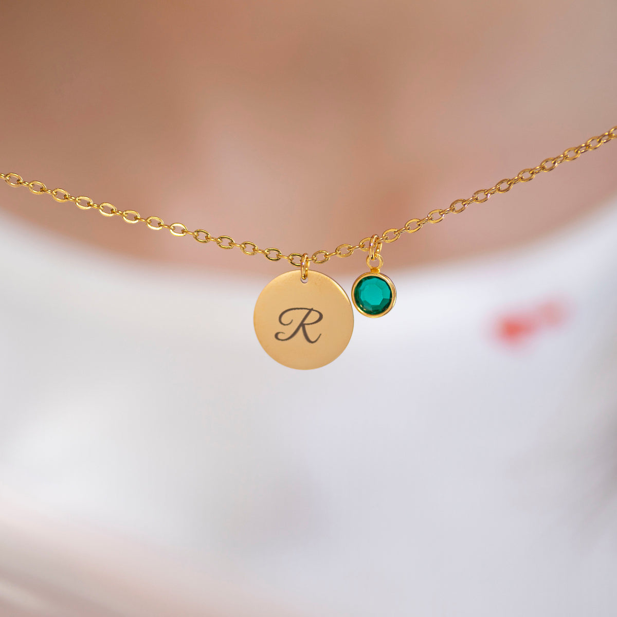Gold script initial birthstone charm necklace with engraved letter R and green birthstone charm on a fine chain against a softly blurred background.