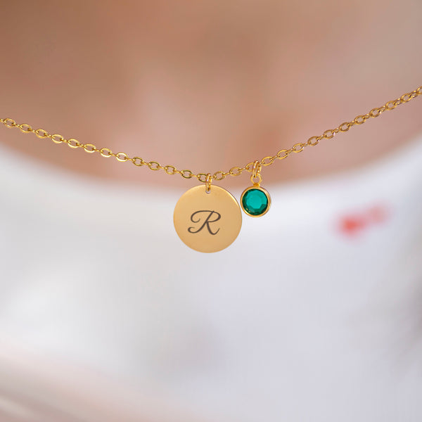 Gold script initial birthstone charm necklace with engraved letter R and green birthstone charm on a fine chain against a softly blurred background.