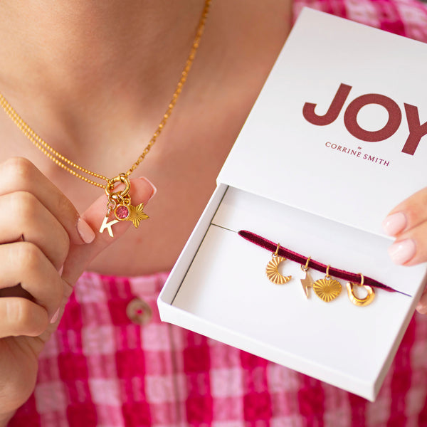 Personalised Week Of Charms Necklace in gold worn with pink birthstone, initial and star charms beside JOY by Corrine Smith gift box with interchangeable charms.
