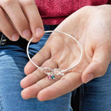 image shows a woman in jeans and a pink jumper holding a silver infinity bangle with birthstone charms