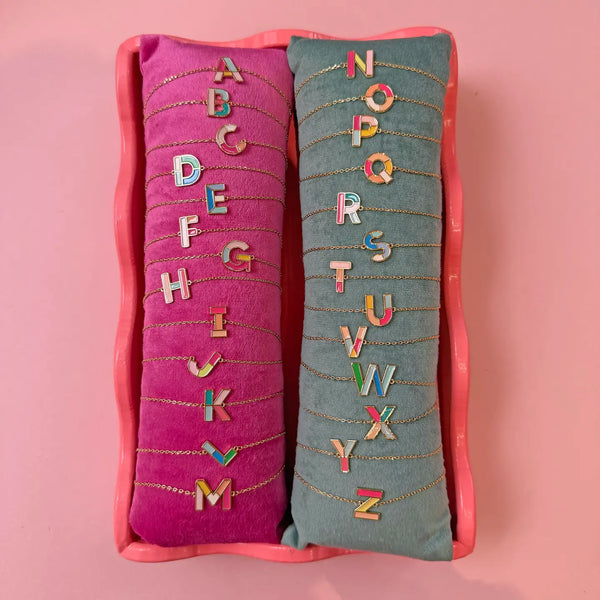 Personalised colourful enamel initial bracelets A to Z with gold plated letter charms displayed on velvet cushions