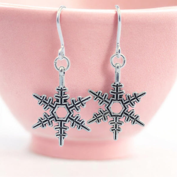 image shows a pair of silver snowflake earrings hanging from a pale pink trinket dish