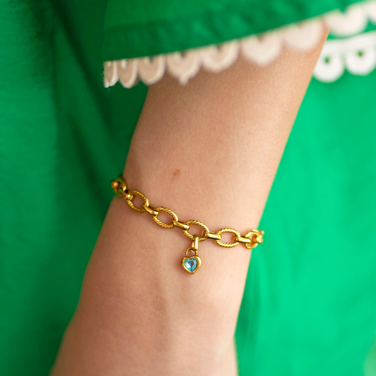 Gold chain bracelet with a blue heart charm displayed on a wrist against a green dress backdrop.