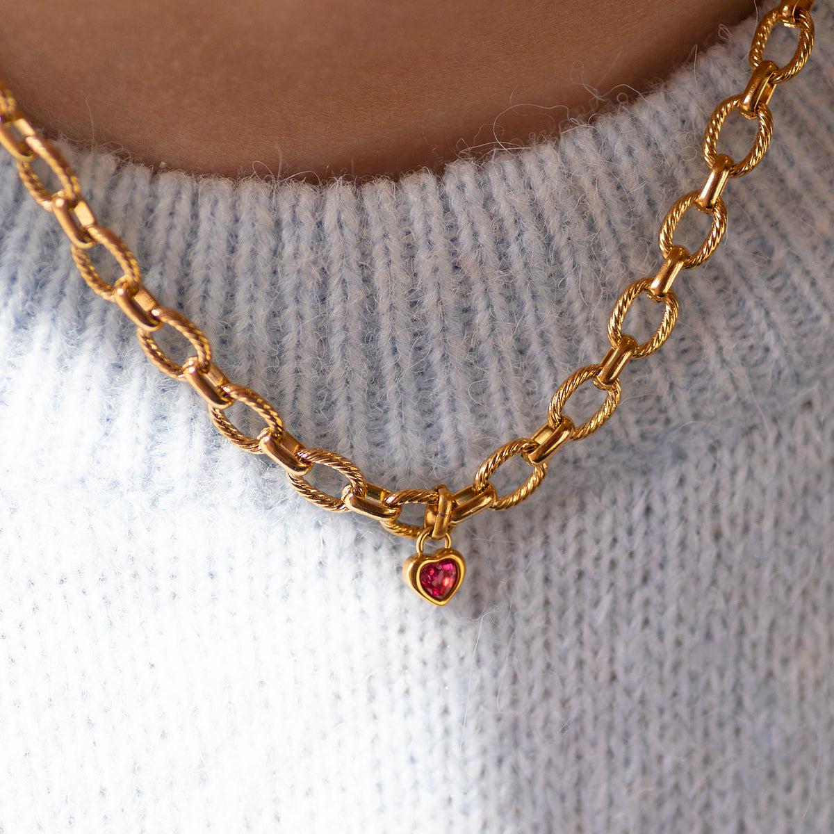 Gold chain necklace with a heart shaped pink gemstone charm on a light grey sweater.