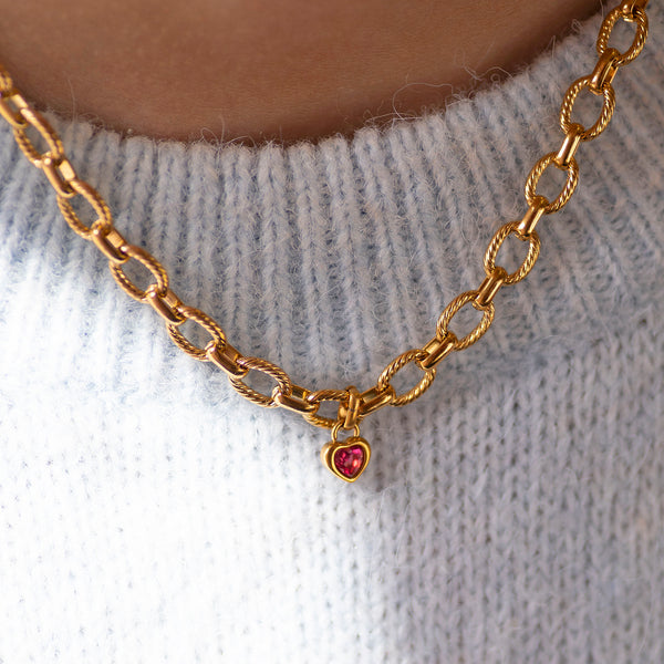 Gold chain necklace with a heart shaped pink gemstone charm on a light grey sweater.