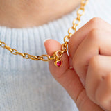 Textured gold chain with a heart shaped red gemstone charm held by a hand against a light background