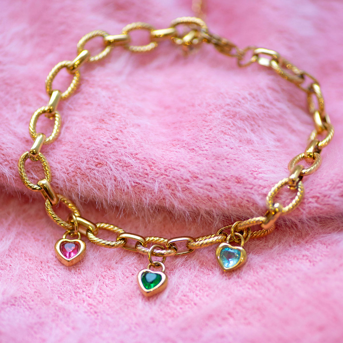 Gold chunky chain bracelet with heart shaped birthstones on a pink fur background