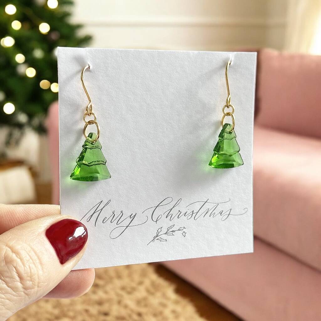 image shows a pair of Christmas tree earrings on a merry christmas card with a christmas living room backgroud