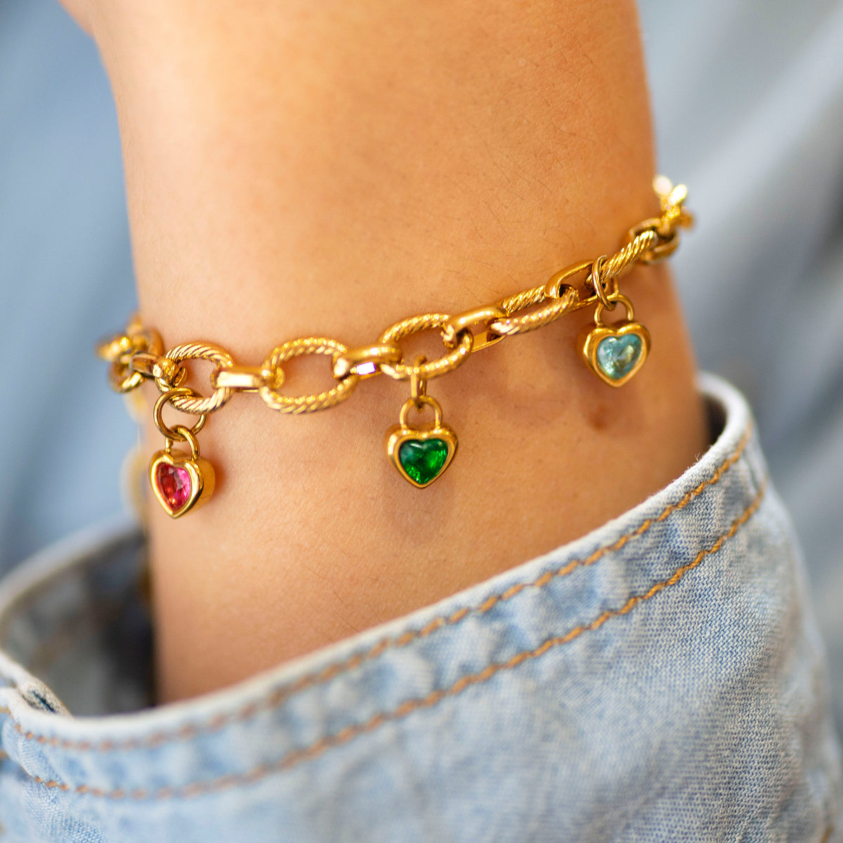 Gold charm bracelet with heart shaped charms on a person's wrist worn with a denim shirt.