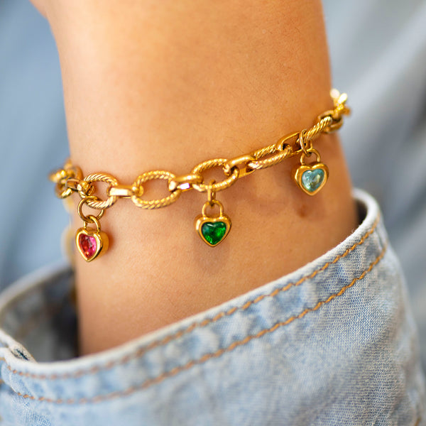 Gold charm bracelet with heart shaped charms on a person's wrist worn with a denim shirt.