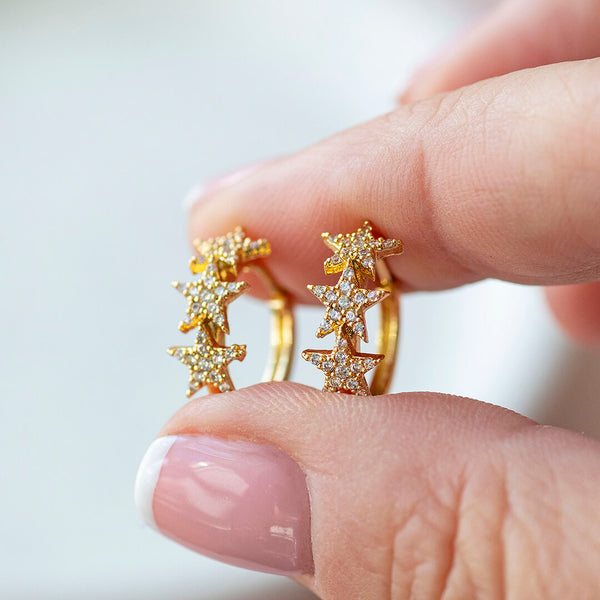 hands holding two gold plated hoops earrings. Each earring has three crystal stars in a row.