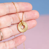 Image shows a gold plated ribbed circle necklace being held by a hand in front of a pink and blue background