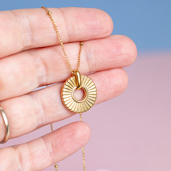 Image shows a gold plated ribbed circle necklace being held by a hand in front of a pink and blue background