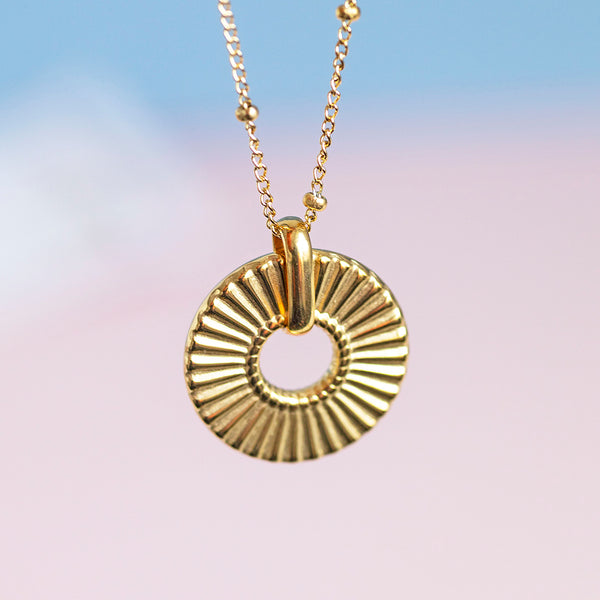 Image shows a gold plated ribbed circle necklace hanging in front of a pink and blue background