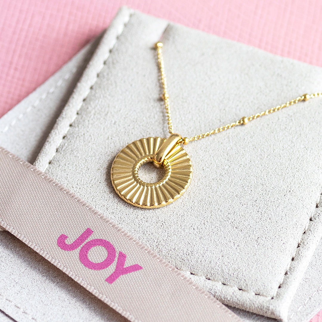 Image shows a gold plated ribbed circle necklace sitting on a suede pouch with the wording joy