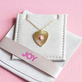 Image shows a gold plated sunburt heart necklace with a pink gemstone at the centre displayed on joy by corrinesmith jewellery pouch