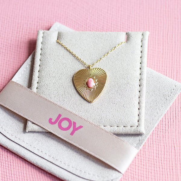 Image shows a gold plated sunburt heart necklace with a pink gemstone at the centre displayed on joy by corrinesmith jewellery pouch